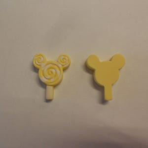 Könnte beinhalten: Zwei gelbe Mickey-Maus-förmige Lollies aus Plastik. Ein Lolli hat ein weißes Spiralmuster.