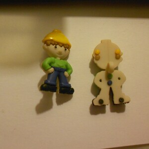 May include: Two plastic figurines, one is a construction worker wearing a yellow hard hat, green shirt, and blue pants, the other is a white figurine with yellow eyes and a blue shirt.