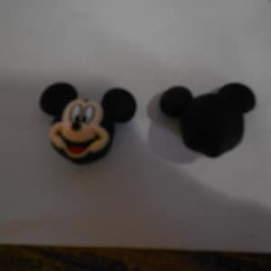 May include: Two Mickey Mouse-themed charms. One charm features Mickey's face with black ears, a tan face, and a red smile. The other charm is a solid black silhouette of Mickey's head. These charms are likely for shoes or accessories.