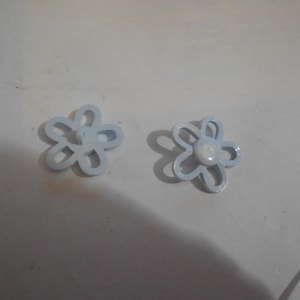 May include: Two light blue flower-shaped buttons. Each button has five petals and a white center. The buttons are made of a smooth material and are set against a white background. These buttons are ideal for sewing and craft projects.