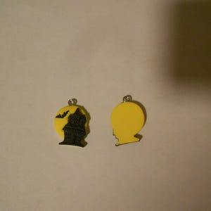 Puede incluir: Dos colgantes de Halloween amarillos. Un colgante presenta una silueta negra de una casa con un murciélago volando por encima. El otro colgante es un círculo amarillo liso.