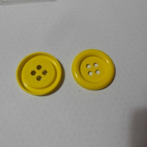 May include: Two bright yellow buttons with four holes each. One button is flat with a slightly raised rim, while the other is a rounded, domed shape. The buttons are made of a smooth, glossy material.