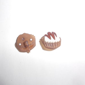 May include: Two brown plastic buttons shaped like cupcakes. One button has white frosting and brown chocolate swirls on top. The other button is plain brown.