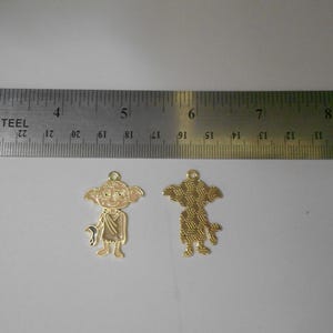 Puede incluir: Dos colgantes metálicos dorados con forma de personaje de dibujos animados, uno con un frente detallado y otro con una parte trasera texturizada. Los colgantes están junto a una regla, midiendo aproximadamente 5 cm de altura.