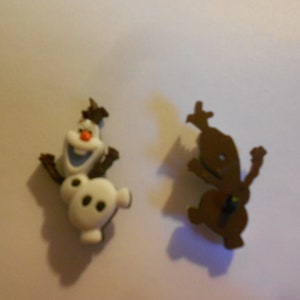May include: Two small plastic figurines, one of Olaf from Frozen, the other of a brown snowman. Both are in a standing position.