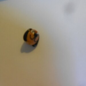 May include: A small, brown ceramic bead with a painted face. The bead has black hair and a mustache.