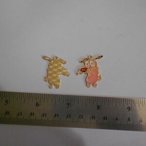May include: Two Courage the Cowardly Dog cartoon character charms. One is gold-colored with a textured pattern, the other is pink with red and black accents. Both have a small loop at the top for attaching to a necklace or bracelet.