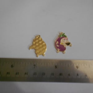 May include: Two Courage the Cowardly Dog enamel charms. One is gold with a textured pattern, the other features Courage in pink, white, and green. A ruler is in the foreground, with measurements in inches.