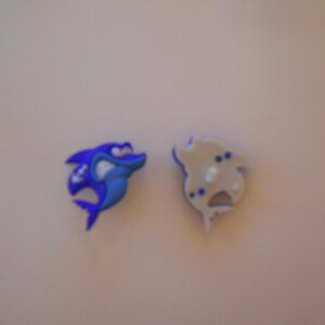 May include: Two cartoon shark-shaped charms. One is blue with white accents and a stern expression. The other is a light tan color with blue and white details. These charms could be used for shoe decoration.