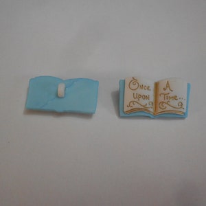 May include: Two light blue book-shaped embellishments. One is closed with a white clasp, the other is open, displaying the words "Once Upon A Time..." in gold script. These are likely for scrapbooking or crafting.
