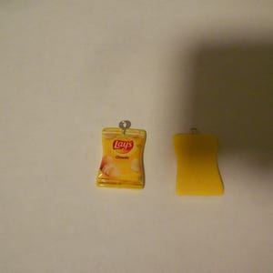 Puede incluir: Dos amuletos amarillos, uno con forma de bolsa de patatas fritas Lay's con el texto "Lay's Classic" y el otro con forma de rectángulo.