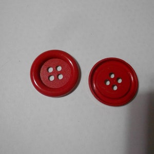 May include: Two red buttons with four holes each, set against a white background. The buttons are circular, with a slightly raised rim and a matte finish. They are likely for sewing or crafts.