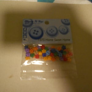 Puede incluir: Una bolsa de plástico transparente llena de botones coloridos. Los botones son de varias formas y colores, incluyendo azul, naranja, morado y verde. La bolsa tiene una cabecera de cartón con el texto "TSTS Home Sweet Home" e imágenes de botones.