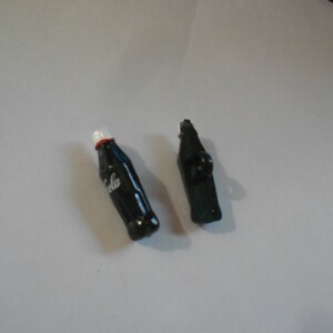 May include: Two black plastic bottle stoppers. One is shaped like a Coca-Cola bottle with the word "Coca-Cola" printed on it. The other is a simple, rectangular shape.