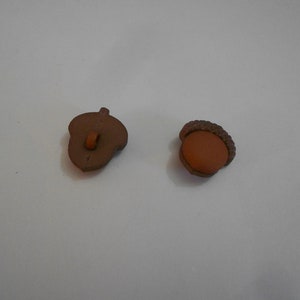以下が含まれることがあります： 茶色のどんぐり型のボタンが2つ。1つのボタンは茶色の帽子、もう1つのボタンは茶色の帽子と茶色のループが付いている。
