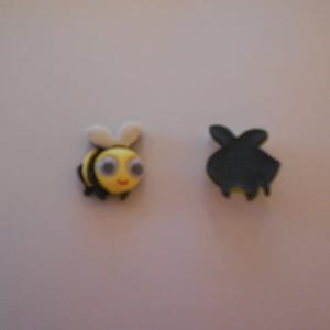 May include: Two small bee-shaped embellishments. One is a yellow bee with black stripes, white wings, and large eyes. The other is a black bee-shaped object with yellow accents. These are likely decorative items.
