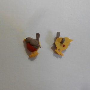 以下が含まれることがあります： ロビン型のプラスチックボタンが2つ。1つのボタンは、茶色の頭、赤い胸、黄色の足を持つロビンの正面を示しています。もう1つのボタンは、黄色のプラスチックでできたロビンの背面を示しています。