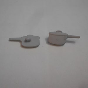 May include: Two white ceramic pot-shaped buttons. One button is a side view of a pot with a handle. The other button is a top view of a pot with a handle.