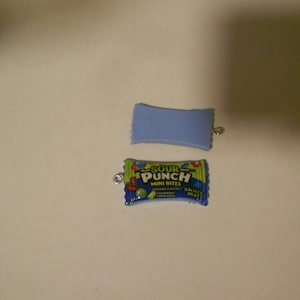 Puede incluir: Dos dijes de plástico azul, uno con forma de barra de caramelo con el texto "Sour Punch Mini Bites" y el otro un simple rectángulo.