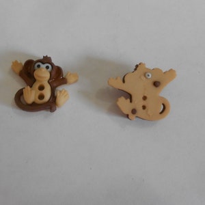 May include: Two plastic buttons shaped like monkeys. The first button is brown with a light brown face and white eyes. The second button is light brown with a brown face and white eyes.