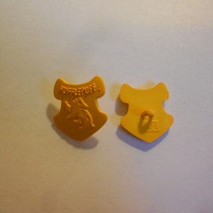 May include: A yellow plastic cookie cutter shaped like the Hufflepuff crest from the Harry Potter series. The front of the cutter features the Hufflepuff badger and the words "Hufflepuff". The back of the cutter has a small hole for hanging.