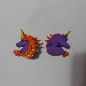 May include: Two purple unicorn buttons with orange glitter and yellow horns. The buttons are approximately 2.5 cm in diameter.