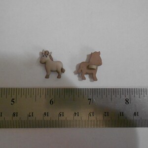 Könnte beinhalten: Zwei kleine, hellbraune Pferdfiguren. Ein Pferd hat eine detaillierte Mähne und einen Schweif, das andere ein schlichtes Design. Ein Metalllineal mit Zentimetermarkierungen liegt im Vordergrund.
