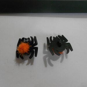 Op de afbeelding: Twee zwarte knoppen in de vorm van een spin met oranje, pluche centra. Eén spin heeft knoopjesogen.