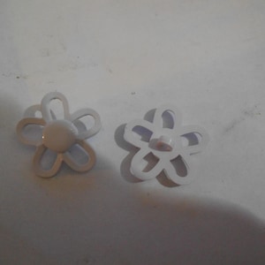 May include: Two white flower-shaped buttons with a central, rounded button. The petals are outlined, and the buttons are set against a white background. These buttons are suitable for sewing and craft projects.
