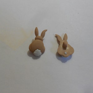 Puede incluir: Dos botones en forma de conejito de color marrón claro. Un botón muestra la parte trasera del conejo con una cola blanca y esponjosa. El otro botón muestra la parte trasera del conejo con un ojal.