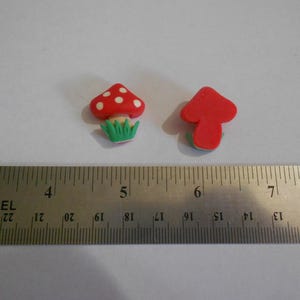 May include: Two small mushroom-shaped figurines. One is red with white spots and green grass, the other is a solid red mushroom. A metal ruler with inch measurements is in the foreground.