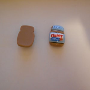 Peut inclure: Deux breloques miniatures sur le thème de la nourriture. L'une est un sandwich au beurre de cacahuète brun, l'autre est un pot de beurre de cacahuète Skippy Creamy avec un couvercle bleu. Le pot porte le nom de la marque en rouge et bleu.