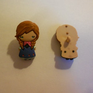 May include: A brown-haired girl with a blue dress and pink bow, a cartoon character button. The back of the button is beige with a hole for sewing.
