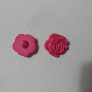 Könnte beinhalten: Zwei rosafarbene Plastikknöpfe in Rosenform. Ein Knopf zeigt die Rückseite mit einem Loch zum Nähen. Der andere Knopf zeigt die Vorderseite mit einem erhabenen Rosendesign.