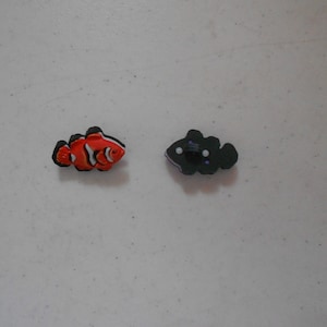 Può includere: Due piccole figurine di pesci di plastica. Una è un pesce pagliaccio arancione e bianco, l'altra è un pesce nero con macchie bianche.