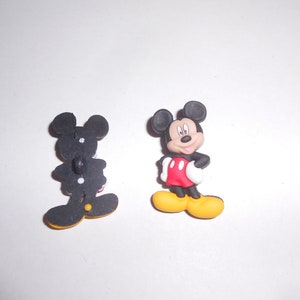 Könnte beinhalten: Zwei Knöpfe zum Thema Micky Maus. Ein Knopf ist eine dreidimensionale Figur von Micky Maus in seinem klassischen Outfit. Der andere Knopf ist eine schwarze Silhouette von Micky Maus mit einem Knopf auf der Rückseite.