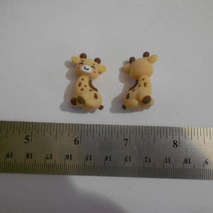 Puede incluir: Dos pequeñas figuras de jirafa sonriendo. Las jirafas son de color canela con manchas y detalles marrones. Una regla metálica está en primer plano, con medidas en centímetros. Las jirafas miden aproximadamente 2,5 cm de altura.