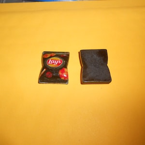 May include: A miniature replica of a Lay's potato chip bag, with the brand name and a depiction of potato chips. The bag is black with red and yellow accents. Next to it is a black rectangular object.