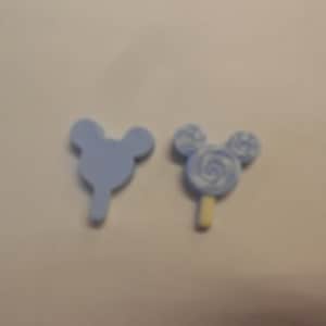 May include: Two light blue Mickey Mouse-shaped embellishments. One is a solid shape, and the other is a lollipop design with a white swirl and a tan stick. These are likely craft supplies or decorative elements.