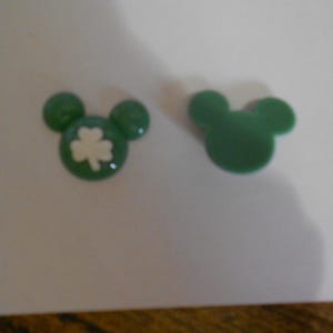 May include: Two green Mickey Mouse head-shaped charms. One charm features a white shamrock design inside the head. The other charm is a solid green Mickey Mouse head. These charms are likely for jewelry or craft projects.