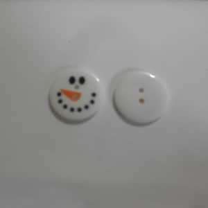 Può includere: Due bottoni in ceramica bianca, uno con un viso di pupazzo di neve nero e arancione e l'altro con due piccoli punti neri.