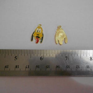 May include: Two gold-colored charms. One charm is a yellow, black, and red Pluto face. The other charm is a gold-colored, textured shape. A metal ruler is in the foreground. The charms are approximately 0.75 inches in length.