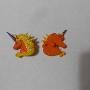 May include: Two orange unicorn buttons with gold glitter and purple horns. One button is facing forward and the other is facing backward.
