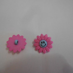 May include: Two pink plastic flower buttons with a smiley face on one. The buttons have a light blue center.