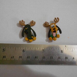 Op de afbeelding: Twee kleine, handgemaakte Pluto-beeldjes met geweien. Eén is groen en zwart, de andere is geel en oranje. Beide hebben zwarte oren en gele voeten. Een metalen liniaal staat op de voorgrond.