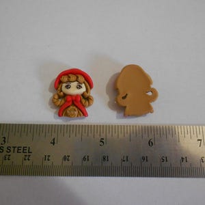 May include: Two small, decorative figurines. One is a detailed, front-facing depiction of a girl in a red hooded cloak. The other is a tan-colored silhouette of the same figure. A stainless steel ruler is in the foreground.