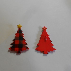 Puede incluir: Dos botones con forma de árbol de Navidad, uno rojo y otro con un patrón de cuadros rojos y negros. Ambos botones tienen una estrella amarilla en la parte superior.