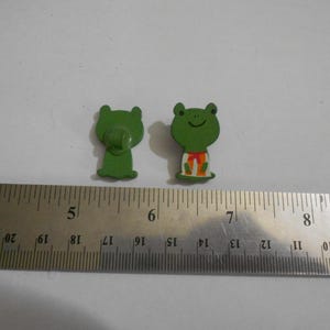 May include: Two green frog-shaped decorative items. One frog has a clip on the back, and the other has a smiling face and a small outfit. A metal ruler with inch measurements is in the foreground.