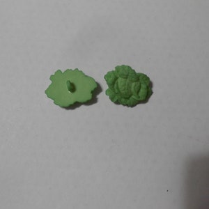 Puede incluir: Dos botones verdes claros con forma de hoja. Un botón muestra la parte posterior con un vástago de botón, y el otro muestra la parte delantera con un diseño de hoja detallado. Los botones son de material liso.