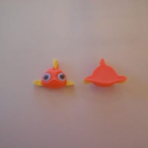 May include: Two orange fish-shaped embellishments with yellow fins and tail. One has googly eyes. The other is a top view of the fish. These are likely craft supplies or decorative elements.